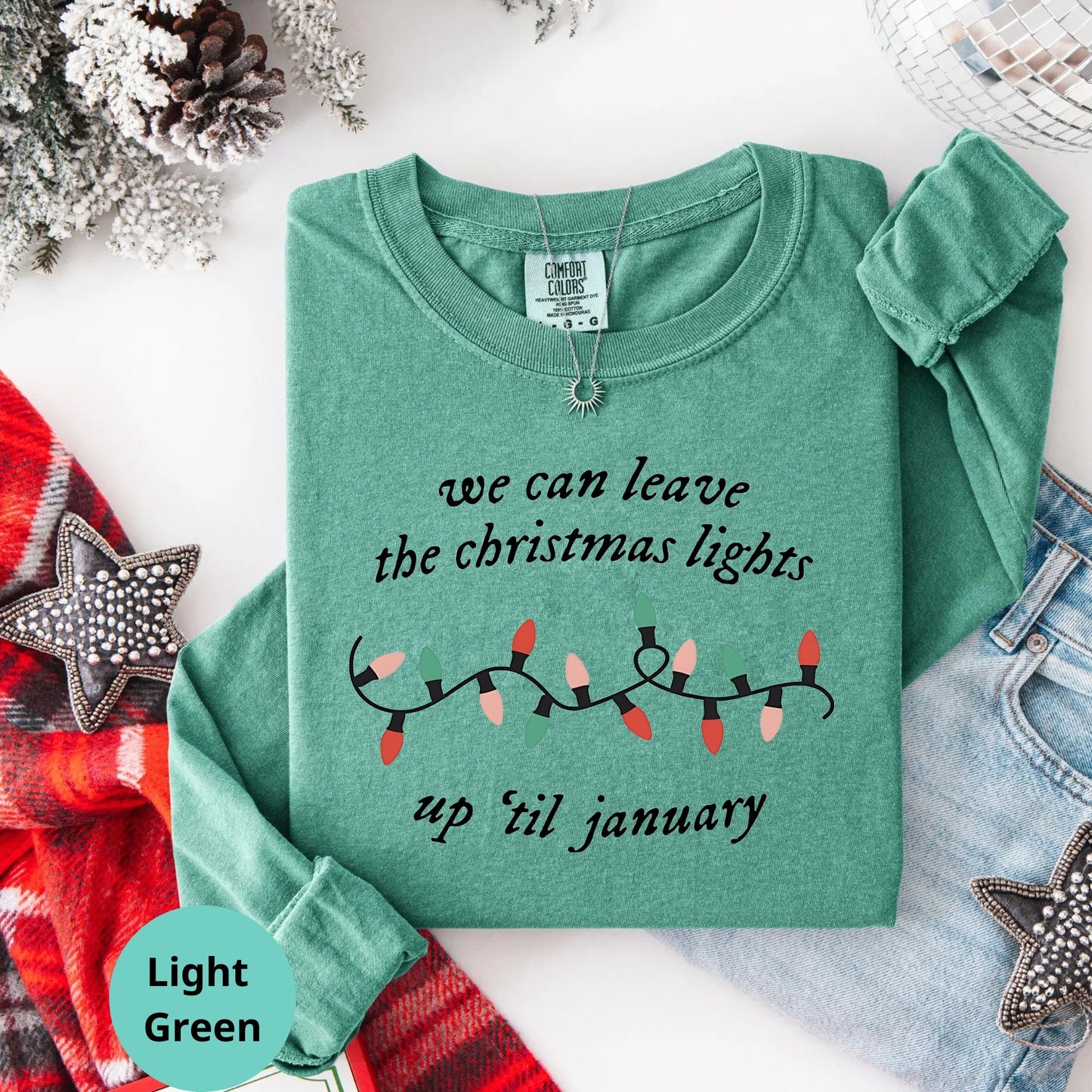 Light green sweatshirt with Christmas lights design and text on a white background with holiday decorations.